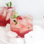 A glass of raspberry cocktail with ice, garnished with mint, sits next to fresh raspberries and a bourbon bottle—a delightful cocktail with raspberries crafted using a classic bourbon drink recipe.