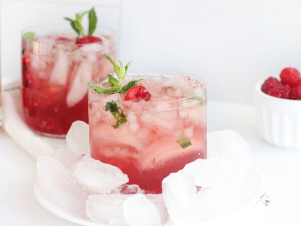 A glass of raspberry cocktail with ice, garnished with mint, sits next to fresh raspberries and a bourbon bottle—a delightful cocktail with raspberries crafted using a classic bourbon drink recipe.