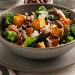 A bowl of Roasted Butternut and Feta Salad featuring greens, feta cheese, roasted pumpkin, and pecans sits on a plate with a fork nearby.