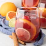 A glass of fruity rum punch sangria with ice, citrus slices, and a cherry sits beside oranges, lime, and cinnamon sticks.