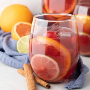 A glass of fruity rum punch sangria with ice, citrus slices, and a cherry sits beside oranges, lime, and cinnamon sticks.