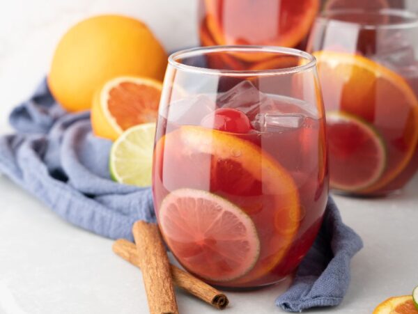 A glass of fruity rum punch sangria with ice, citrus slices, and a cherry sits beside oranges, lime, and cinnamon sticks.
