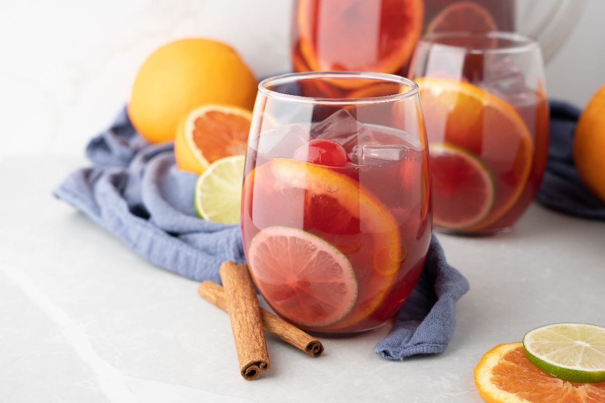A glass of Pinot noir fruit sangria with ice, citrus slices, and cherries sits next to cinnamon sticks and fresh fruit.