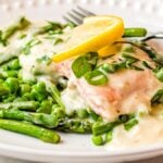 Salmon with white wine sauce, creamy asparagus, peas, and lemon slices on a white plate with a fork—an easy salmon recipe perfect for dinner.