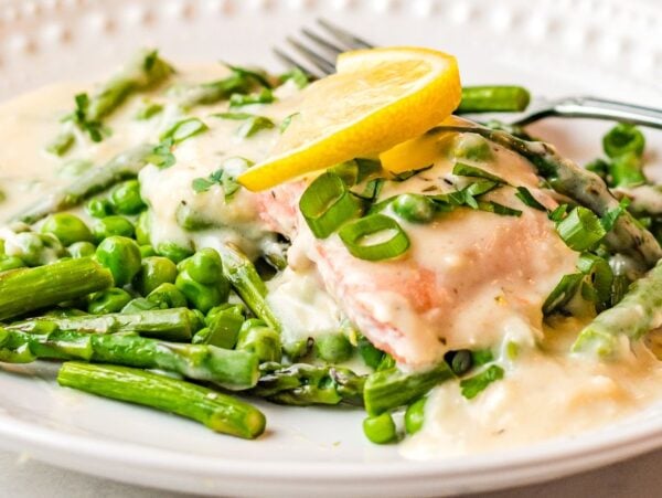 Salmon with white wine sauce, creamy asparagus, peas, and lemon slices on a white plate with a fork—an easy salmon recipe perfect for dinner.