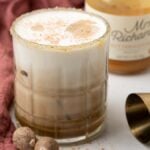 Iced coffee with frothy milk in a glass, using butterscotch schnapps and coffee liqueur, garnished with spices, sits next to a bottle and jigger on a pink cloth—a tasty fall cocktail perfect for cozy moments.