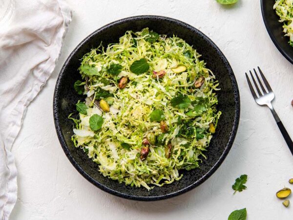 Black bowl of shredded green salad with shaved brussels sprouts, mint, pistachios, and cheese on a white surface, next to a fork.