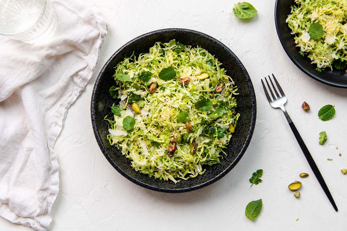 A black bowl of shredded green salad with herbs and pistachios, featuring shaved brussels sprouts, sits beside a fork and a white napkin—a refreshing summer salad recipe.