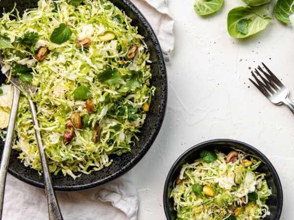 Shaved brussels sprouts and herbs tossed with pistachios make this vibrant Brussels sprouts salad recipe, served in black bowls on a white table with forks—a perfect summer salad recipe.