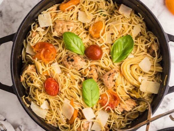 A pot of Sheet Pan Chicken Caprese Pasta with cherry tomatoes, basil leaves, and shaved parmesan on a marble countertop—an easy chicken dinner perfect for any night.