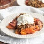 A serving of fruit cobbler topped with whipped cream on a white plate, made with fresh strawberries and peaches—an easy summer dessert that's perfect for any occasion.