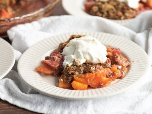 A serving of fruit cobbler topped with whipped cream on a white plate, made with fresh strawberries and peaches—an easy summer dessert that's perfect for any occasion.