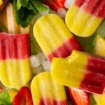 Colorful, refreshing ice pops with red and yellow layers, surrounded by ice, strawberries, pineapple slices, and a hint of mint—an easy layered popsicle recipe perfect for summer treats.