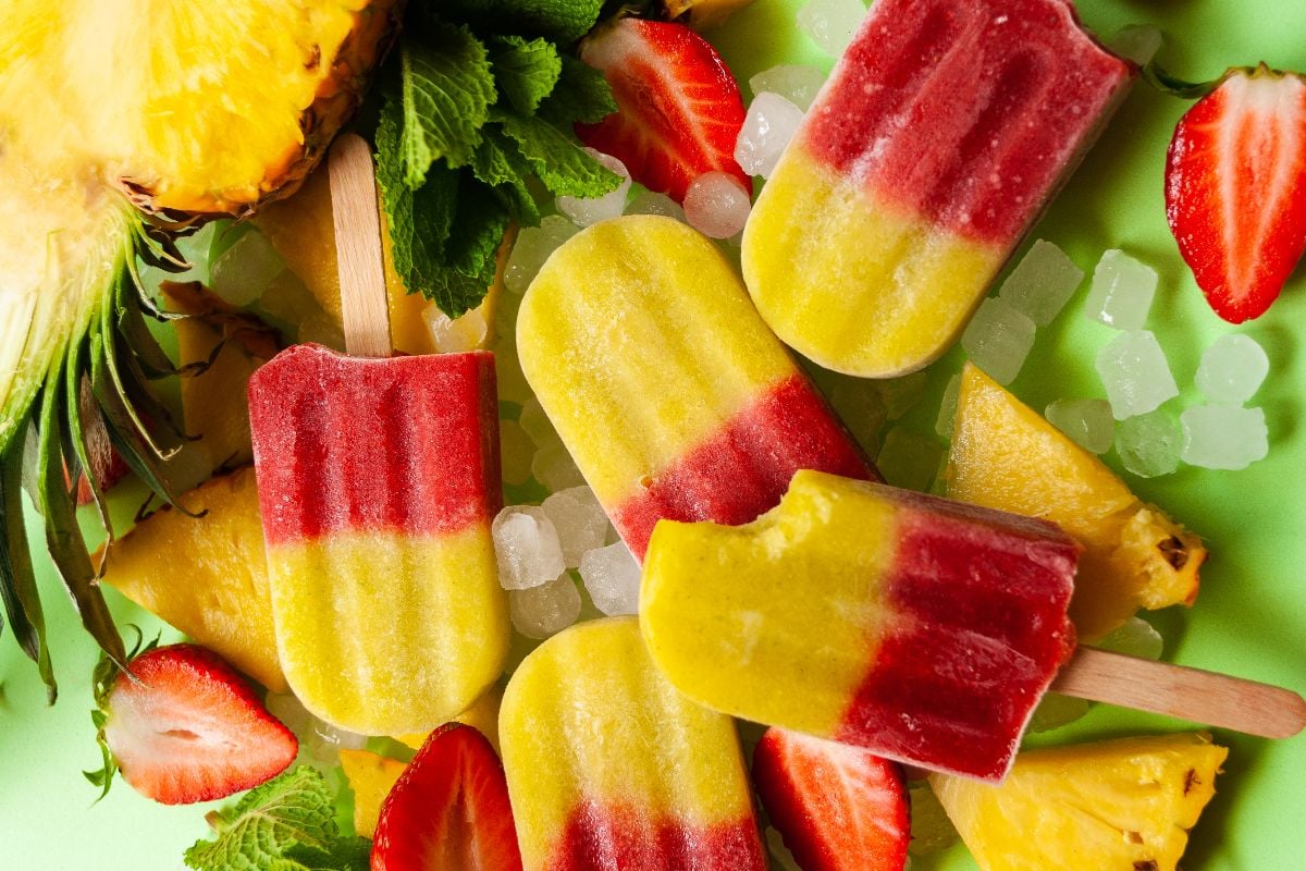 Refreshing Strawberry Pineapple Popsicles with ice, pineapple slices, and strawberries on a green background—an easy layered popsicle recipe bursting with fresh fruit flavors.