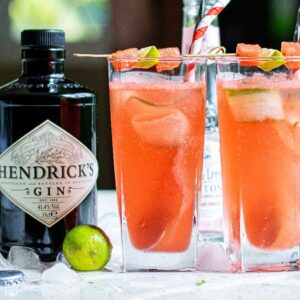 Two tall glasses of pink cocktails with cut cucumber ribbons and fruit skewers stand beside a Hendrick's gin bottle and lime, perfect for a refreshing Summer gin and tonic recipe.