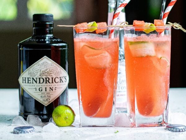 Two tall glasses of pink cocktails with cut cucumber ribbons and fruit skewers stand beside a Hendrick's gin bottle and lime, perfect for a refreshing Summer gin and tonic recipe.