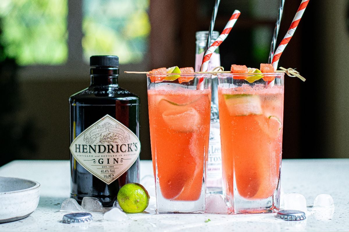Two tall glasses of pink Watermelon Gin Tonic with lime and cut cucumber ribbons sit next to a Hendrick’s gin bottle and ice on a white surface—your new favorite summer gin and tonic recipe.
