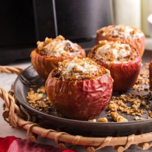 Baked apples stuffed with oats and nuts on a black plate, drizzled with maple syrup, make an easy air fryer apple dessert served on a woven tray.