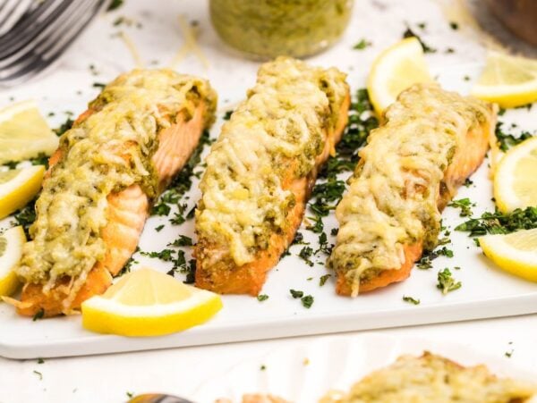 Baked salmon fillets topped with pesto and melted cheese, inspired by Air Fryer Parmesan Pesto Salmon, garnished with lemon wedges and herbs on a white plate.