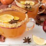 Glass mugs of spiced apple cider with lemon slices, star anise, and cinnamon sticks surround apples in this cozy Apple Cider Fireball Hot Toddy.