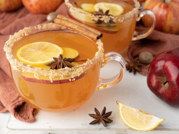 Glass mugs of spiced apple cider with lemon slices, star anise, and cinnamon sticks surround apples in this cozy Apple Cider Fireball Hot Toddy.