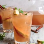 Two glasses of cold apple cider and rum with lime, mint, and cinnamon sticks sit next to a pitcher on a marble surface.