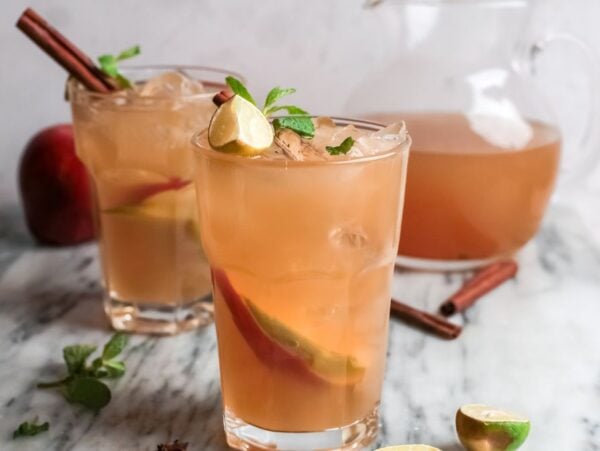 Two glasses of cold apple cider and rum with lime, mint, and cinnamon sticks sit next to a pitcher on a marble surface.
