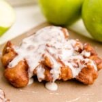 Apple Cinnamon Fritters with a sweet white glaze rest on brown paper, surrounded by whole and sliced green apples—an irresistible take on classic fried apple donuts.