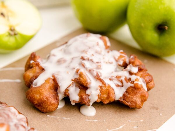 Apple Cinnamon Fritters with a sweet white glaze rest on brown paper, surrounded by whole and sliced green apples—an irresistible take on classic fried apple donuts.