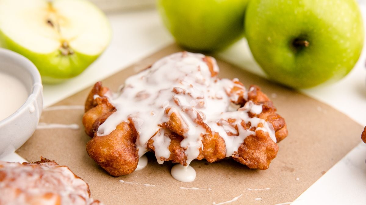 Apple Cinnamon Fritters with a sweet white glaze rest on brown paper, surrounded by whole and sliced green apples—an irresistible take on classic fried apple donuts.
