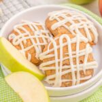 Three Apple Pie Muffin Top Cookies with Vanilla Maple Glaze sit on a white plate beside two apple slices, set atop a green checkered cloth.
