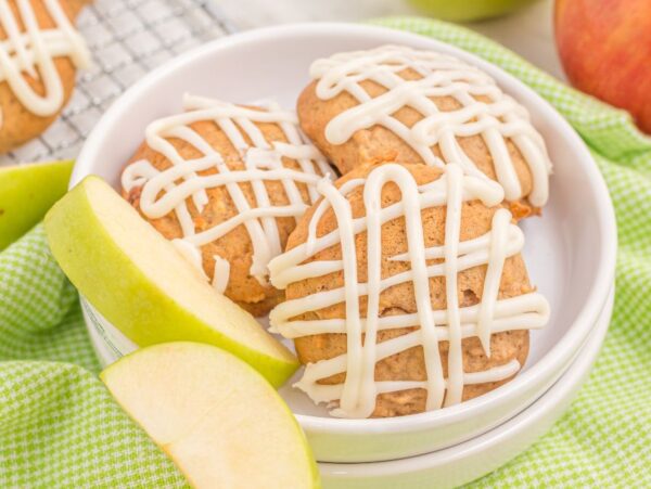 Three Apple Pie Muffin Top Cookies with Vanilla Maple Glaze sit on a white plate beside two apple slices, set atop a green checkered cloth.