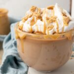 A glass mug of creamy coffee topped with whipped cream, caramel drizzle, and cookie crumbles—perfect for a hot peanut butter drink or as inspiration for a Boozy Salted Peanut Butter Hot Chocolate.