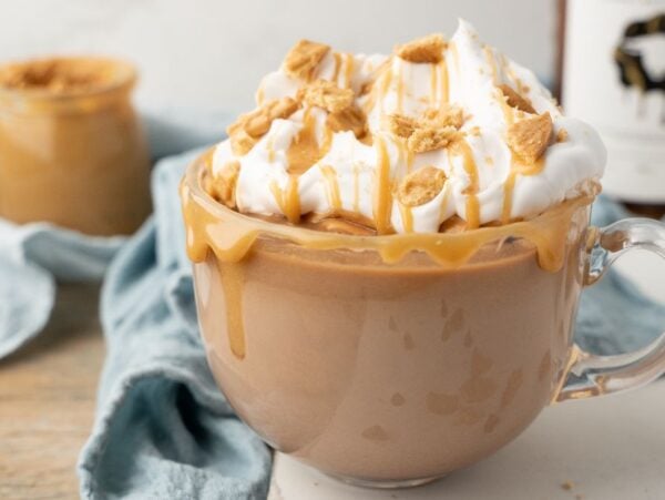 A glass mug of creamy coffee topped with whipped cream, caramel drizzle, and cookie crumbles—perfect for a hot peanut butter drink or as inspiration for a Boozy Salted Peanut Butter Hot Chocolate.
