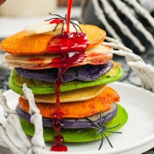 A stack of colorful pancakes with red syrup, topped with fake spiders and surrounded by skeleton hands—perfect for spooky breakfast ideas at your next Halloween party food spread.