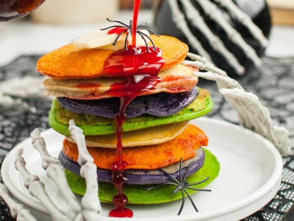 A stack of colorful pancakes with red syrup, topped with fake spiders and surrounded by skeleton hands—perfect for spooky breakfast ideas at your next Halloween party food spread.