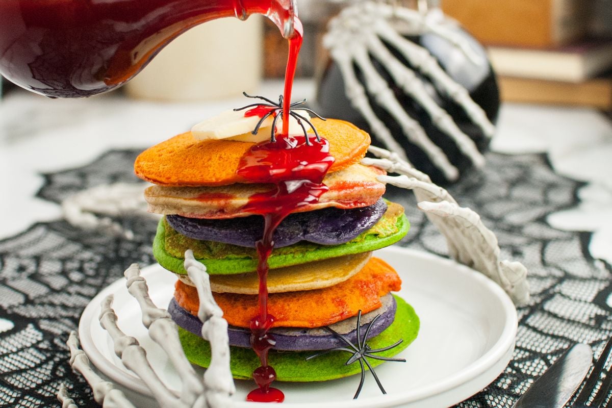 Colorful Halloween Pancakes topped with butter and red syrup are held by skeleton hands with fake spiders—perfect for spooky breakfast ideas and fun Halloween recipes for kids.