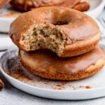 Two Gluten Free Cinnamon Spice Donuts on a plate, one with a bite taken out, rest next to cinnamon sticks—a tempting treat for fans of gluten free baked goods.