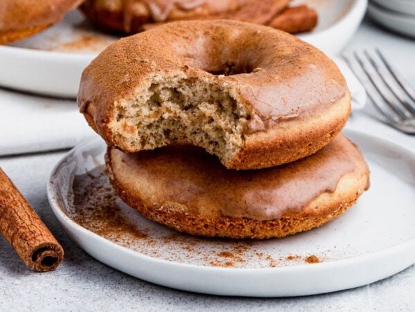 Two Gluten Free Cinnamon Spice Donuts on a plate, one with a bite taken out, rest next to cinnamon sticks—a tempting treat for fans of gluten free baked goods.