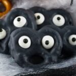 Black Halloween Monster Eye Pretzels decorated with candy eyes, arranged on a spooky cobweb background—an eerie and fun Halloween snack for kids.