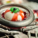 A creepy eyeball dessert in a metal dish held by a skeleton hand, with a plastic spider on the rim—perfect for gross looking Halloween food and spooky Halloween party food ideas. This treat is also a fun gluten free Halloween dessert option.
