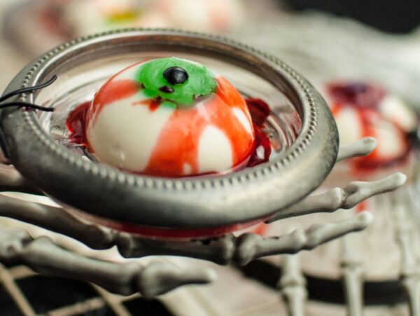 A creepy eyeball dessert in a metal dish held by a skeleton hand, with a plastic spider on the rim—perfect for gross looking Halloween food and spooky Halloween party food ideas. This treat is also a fun gluten free Halloween dessert option.