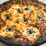 Pizza with black charcoal crust, made from charcoal pizza dough, topped with cheese, pepperoni, and pizza with black olive spiders—perfect for Halloween pizza ideas. Served on a round baking tray.