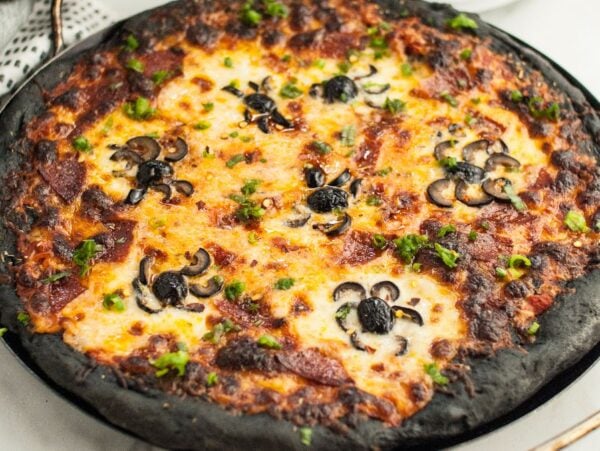 Pizza with black charcoal crust, made from charcoal pizza dough, topped with cheese, pepperoni, and pizza with black olive spiders—perfect for Halloween pizza ideas. Served on a round baking tray.