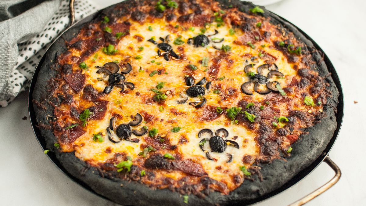 Pizza with black charcoal crust, made from charcoal pizza dough, topped with cheese, pepperoni, and pizza with black olive spiders&mdash;perfect for Halloween pizza ideas. Served on a round baking tray.
