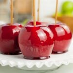 Four shiny red homemade candy apples with sticks rest on a white glass plate, with a blurred kitchen background.