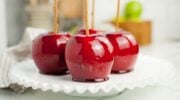 Four shiny red homemade candy apples with sticks rest on a white glass plate, with a blurred kitchen background.