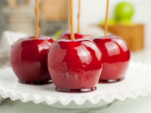 Four shiny red homemade candy apples with sticks rest on a white glass plate, with a blurred kitchen background.