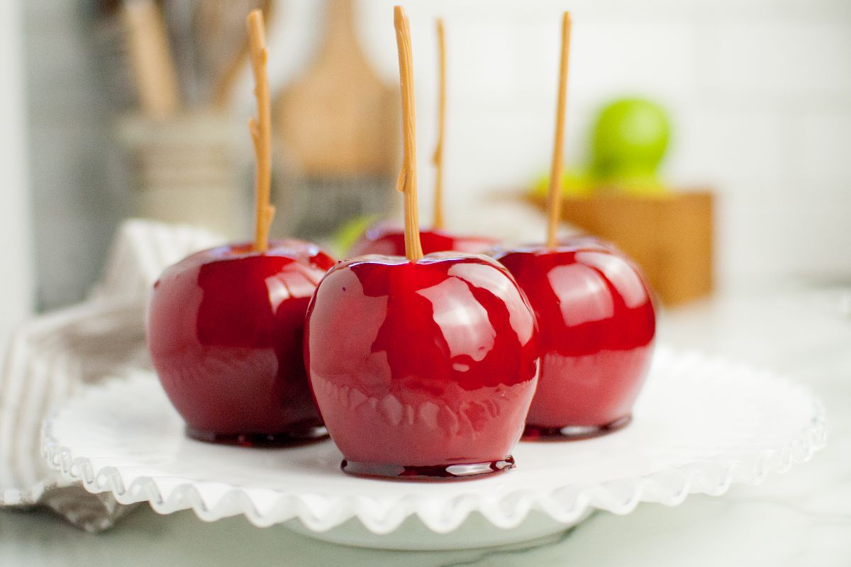 Four shiny red homemade candy apples with wooden sticks on a white ruffled plate, with a blurred kitchen background—perfect inspiration if you want to learn how to make candy apples on a stick at home.