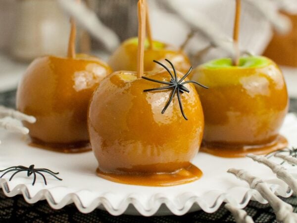 Homemade caramel coated apples with plastic spiders sit on a white plate, surrounded by fake skeleton hands—these are some of the best caramel apples for your spooky party.